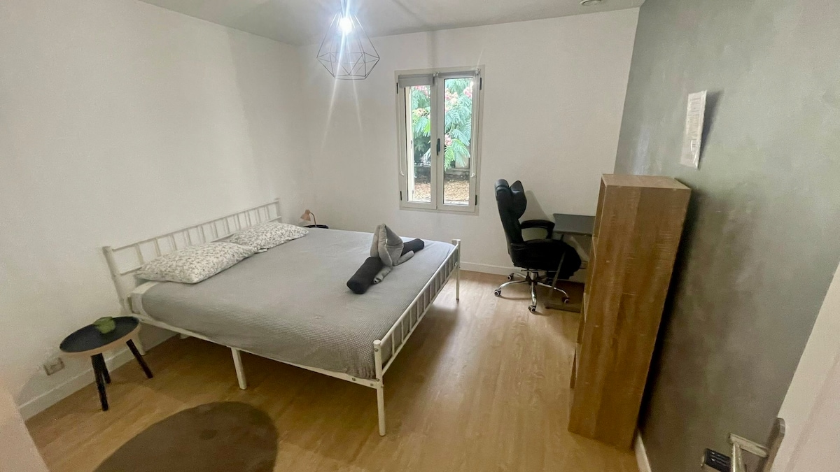 A tranquil bedroom is highlighted by a spacious bed adorned with soft linens. A minimalist desk chair is positioned beside a wooden desk. Natural light filters through the window, offering a view of the greenery outside. A comfortable rug is placed on the floor.