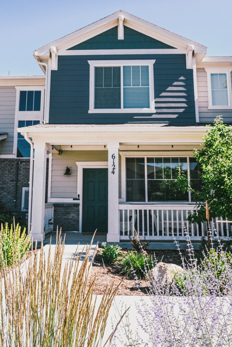 The townhome exterior features a modern design with blue siding and white trim. A welcoming front porch is visible, flanked by lush green plants and lavender. Natural light reflects off the window panes, enhancing the inviting entrance.