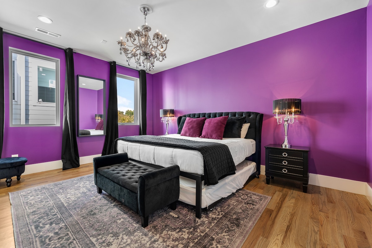 Chumba cha kulala chenye ujasiri na kifahari chenye kuta za zambarau, shada za taa za kifahari na matandiko ya kifahari, kinafaa kwa usiku wa kupumzika katika mtindo mzuri.