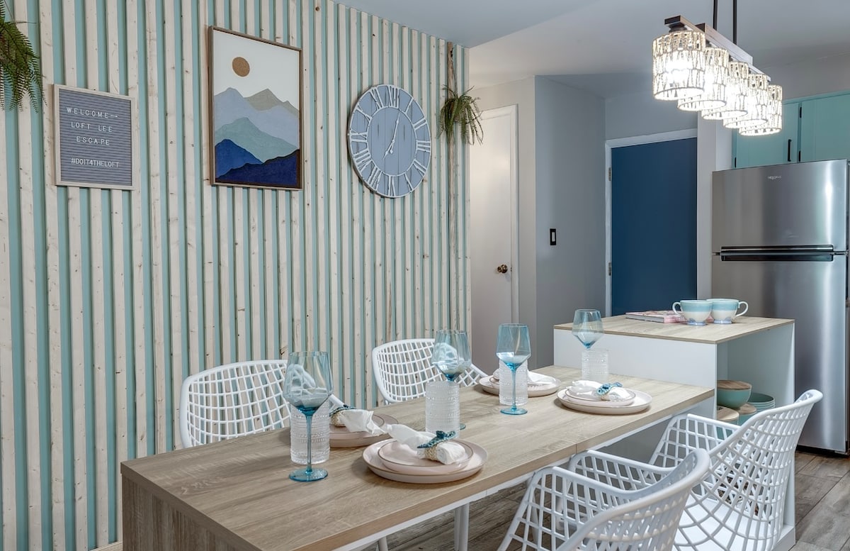 A modern dining area features a long wooden table set for six, surrounded by white, woven chairs. A large clock adorns the blue-striped wall, complemented by framed artwork depicting mountains. Soft lighting is provided by unique pendant lights above the table.