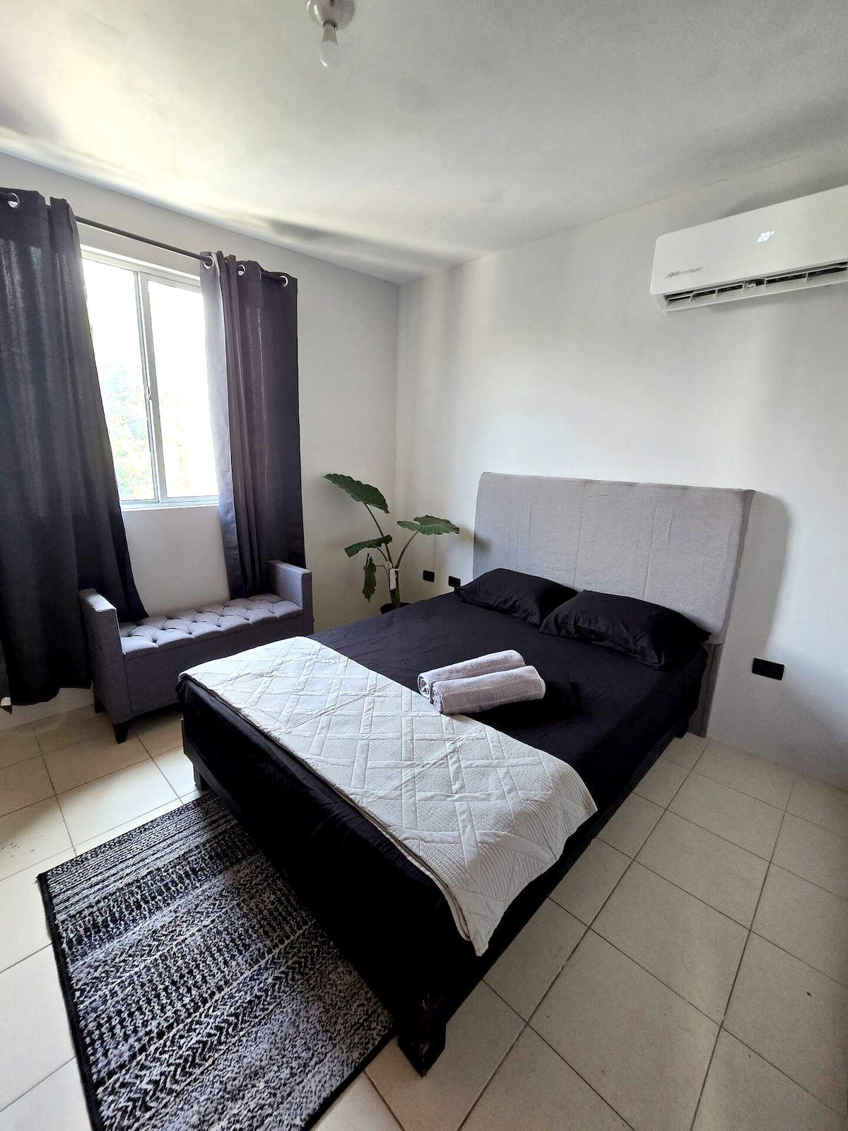 A well-lit sleeping area features a spacious bed with dark bedding and a grey upholstered headboard. A chair is positioned next to a window adorned with blackout curtains. A plant sits near the window, adding a touch of greenery to the neutral-colored room.