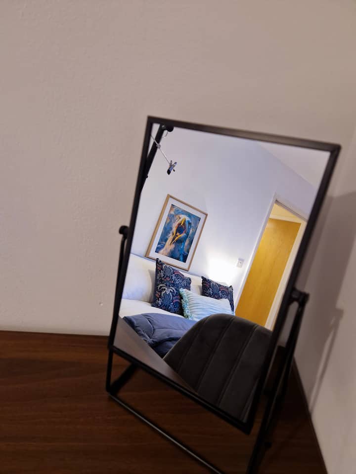 Este acogedor dormitorio cuenta con una cama de tonos suaves vestida con almohadas oscuras estampadas y un cojín a rayas. Un espejo de madera refleja la serenidad del espacio, las obras de arte enmarcadas sobre la cama aportan personalidad y una cálida puerta de madera completa el acogedor ambiente.