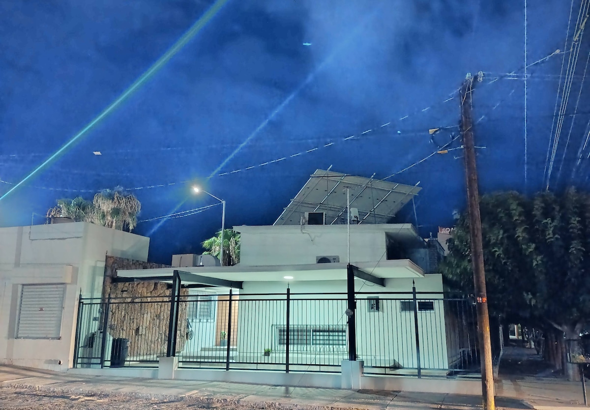 A modern, single-story home is shown at night, illuminated by street lamps. The building features a minimalist design with a fence enclosing a front patio. Solar panels are mounted on the roof, and tall trees provide a natural backdrop.