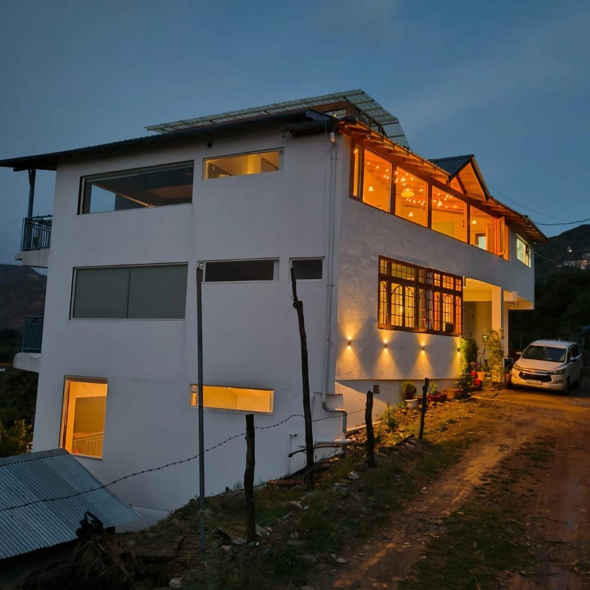 A modern house is showcased at dusk, featuring large windows that offer a glimpse of warm interior lighting. The structure sits on uneven terrain, with outdoor spaces illuminated by soft lighting, and a parked vehicle is visible at the side.