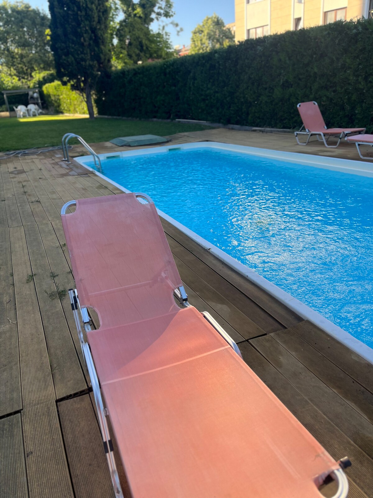 A swimming pool is positioned within a wooden deck area, complemented by a soft pink lounge chair nearby. Lush green hedges surround the space, creating a secluded atmosphere. The clear blue water of the pool reflects sunlight, inviting relaxation in a tranquil outdoor setting.