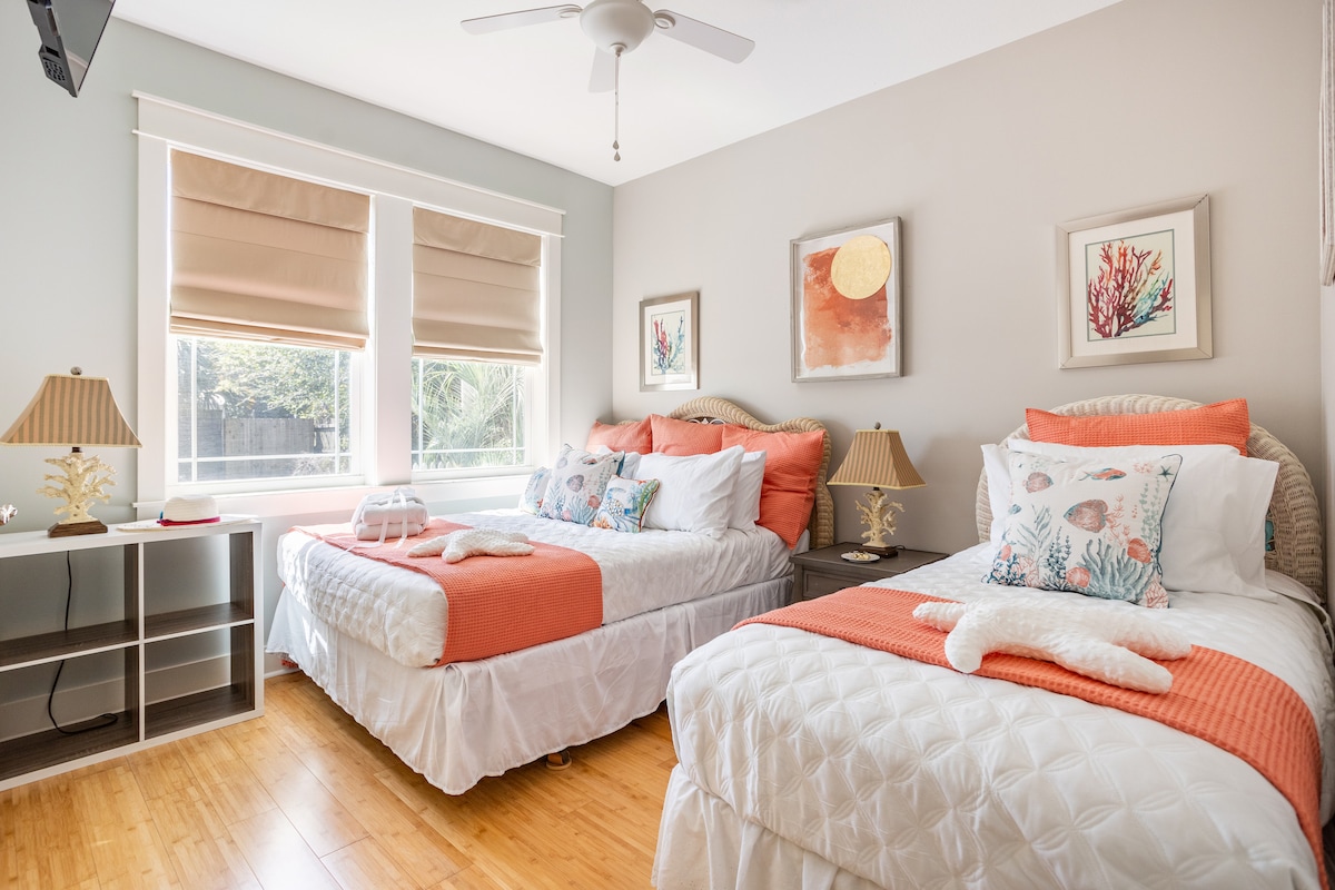 A bright bedroom features two beds, one with a coral-colored throw blanket and decorative pillows. Nightstands with lamps are placed beside each bed, and large windows allow natural light to fill the space, highlighting the neutral decor and soft textures throughout.