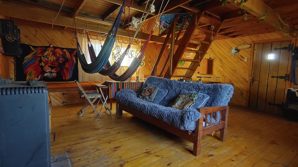 A wooden interior space features a cozy sofa covered with soft blue fabric. Hammocks are suspended overhead, adding a relaxed ambiance. Colorful artwork adorns the walls, including a large lion painting. A dining table and foldable chairs are positioned nearby, providing a functional area.
