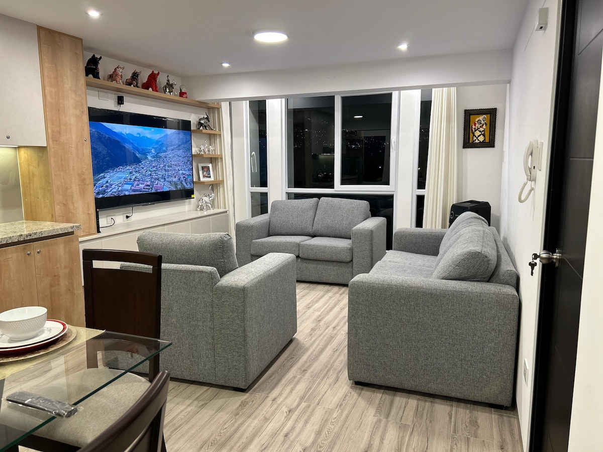 A modern living area features a gray sectional sofa and two armchairs, arranged for conversation. A large window offers natural light and a view of the city. A flat-screen television is mounted on the wall, and a dining table is visible in the corner.