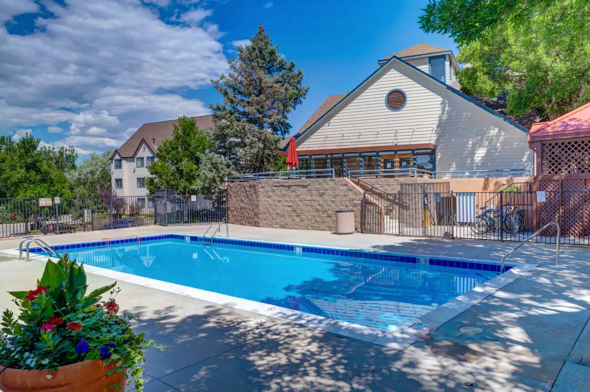 A community pool area is presented, featuring a clear blue swimming pool surrounded by a concrete deck. Lush greenery and colorful flowers are seen in planters nearby. Additional seating and a shaded area are visible in the background, offering a pleasant outdoor space.