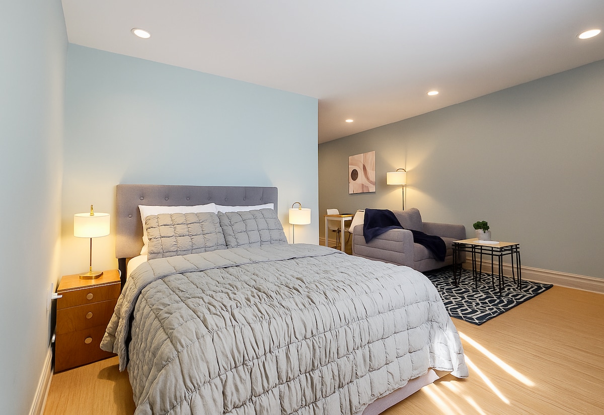 A spacious bedroom features a plush Queen-size bed draped in a soft quilt, paired with bedside lamps for gentle lighting. A comfy loveseat and an area rug add texture to the tranquil space, which includes a table in the background, enhancing the cozy atmosphere.