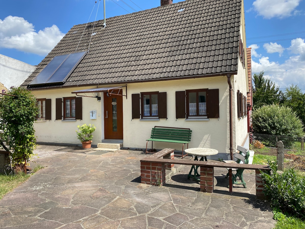 A charming countryside house is presented with a sloped roof and contrasting wooden shutters. A small patio area is visible, featuring a round table and benches. Surrounding greenery complements the inviting exterior, with clear skies adding to the serene atmosphere.
