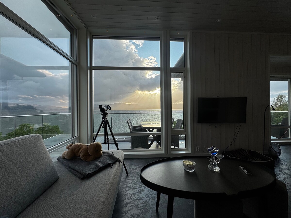 A spacious living area features large windows providing views of the sea and surrounding landscape. A telescope is positioned nearby for observation. Soft furnishings include a cozy gray couch draped with a blanket and a round table topped with a decorative bowl and a drink.