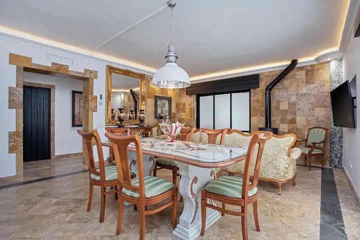 A dining area features elegantly carved wooden chairs surrounding a round table, complemented by a decorative centerpiece. A cozy sitting area is visible in the background, highlighted by natural stone walls and soft lighting, enhancing the room's inviting ambiance.