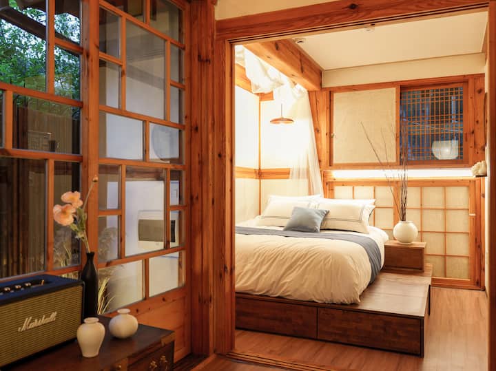 До стъклената врата с бамбукови огради и сини растения има удобна спалня в хотелски стил. Има удобно легло с чисти одеяла и пухкави възглавници, а таванът е покрит с бял балдахин, което създава усещане за хотел. Традиционните решетъчни прозорци зад леглото придават елегантност, а белият порцелан на нощното шкафче и традиционната скрина във всекидневната пред спалнята придават сантименталност с високоговорителите Marshall и малките вази. Това е хотелска спалня с удобно и пухкаво легло за уютна почивка.