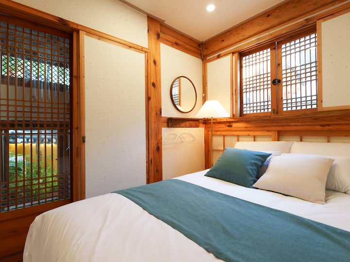 Синя копринена покривка и възглавници са удобно поставени върху чисто и пухкаво легло, а традиционен прозорец на ханок, дървена рамка и кръгло огледало добавят нотка на елегантност към помещението. Слънчевата светлина се разпространява нежно, създавайки уютна и емоционална атмосфера.