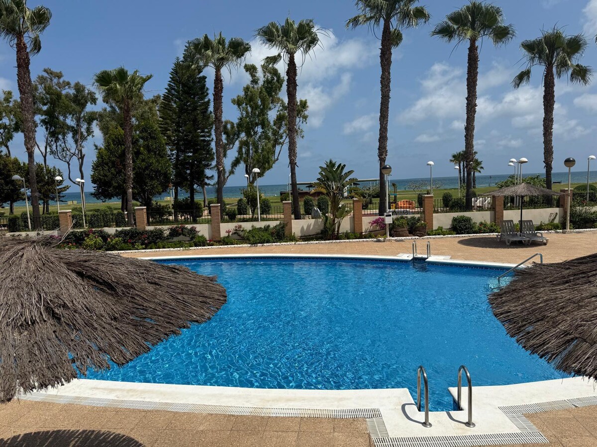 A clear blue swimming pool is surrounded by palm trees and landscaped gardens. Thatched umbrellas provide shade over the poolside area. In the background, the ocean is visible, complementing the tranquil outdoor environment.