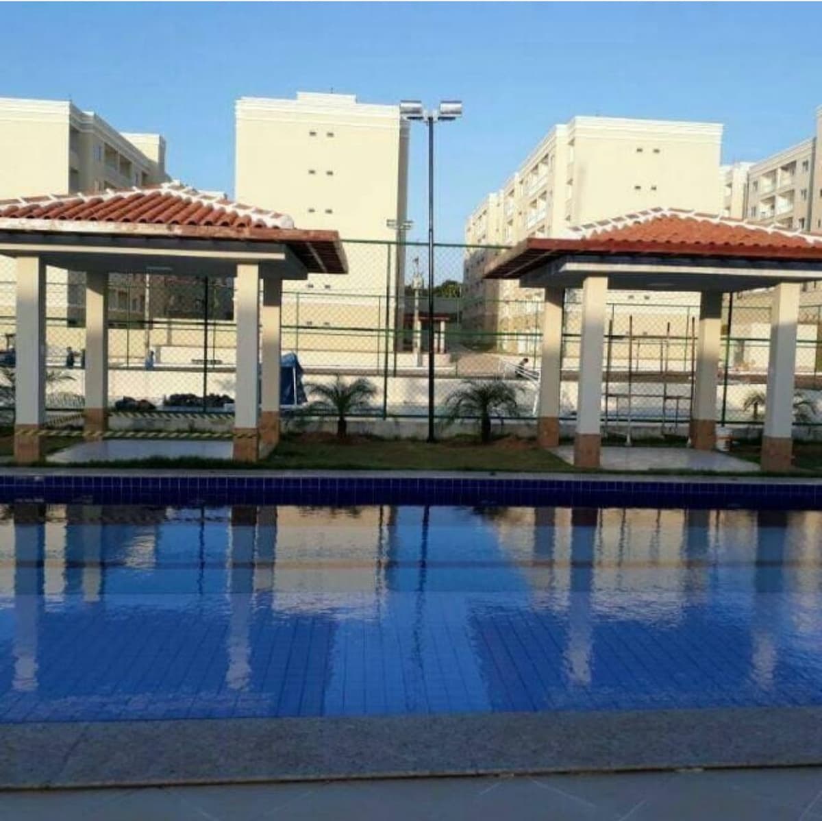 A swimming pool is framed by two covered areas featuring sloped roofs. The clear blue water reflects the surrounding structures and sky. In the background, a sports court is visible, along with landscaped greenery, contributing to a relaxed outdoor environment.