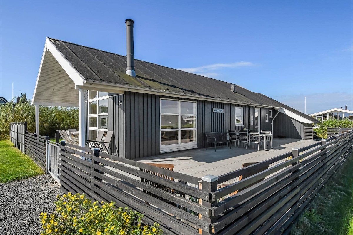 A modern holiday home is presented, featuring a sloped roof and large windows that provide ample natural light. A spacious patio area is visible, equipped with outdoor seating. The surrounding yard is fully fenced, and greenery can be seen in the vicinity.