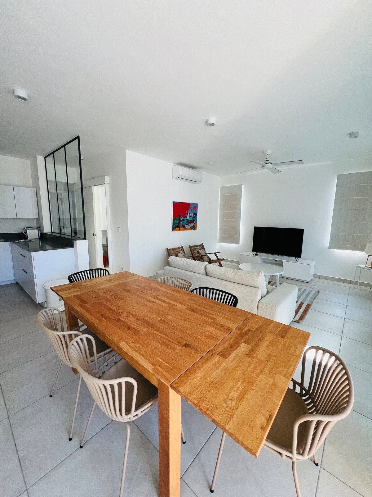 An open living area features a wooden dining table surrounded by six modern chairs, with a cozy seating area in the background. A flat-screen TV is mounted on a light wall, while large windows provide ample natural light, enhancing the spacious atmosphere.