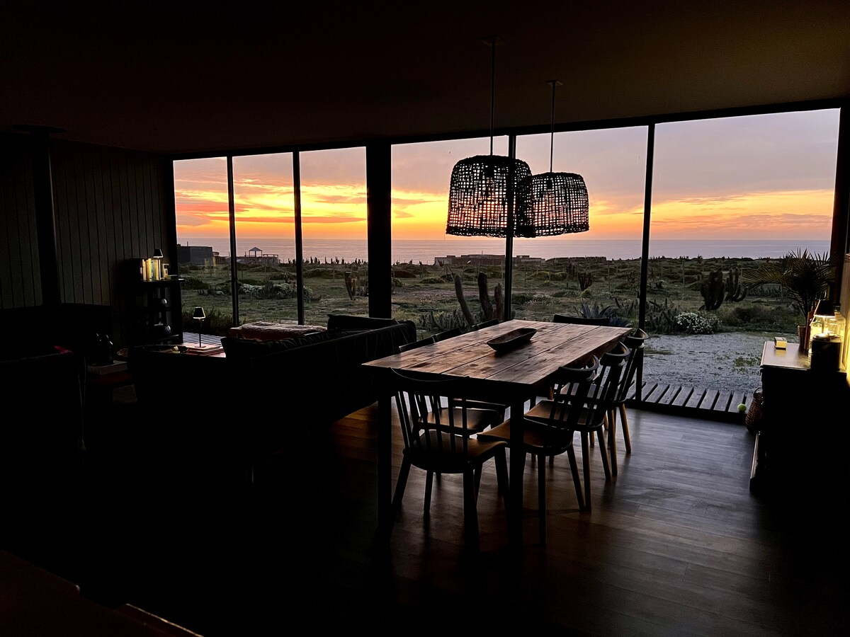 A dining area is featured with a large wooden table surrounded by several chairs. Floor-to-ceiling windows offer a view of a sunset, illuminating the space with warm light. The scene outside includes natural landscapes and distant structures, enhancing the tranquil ambiance.