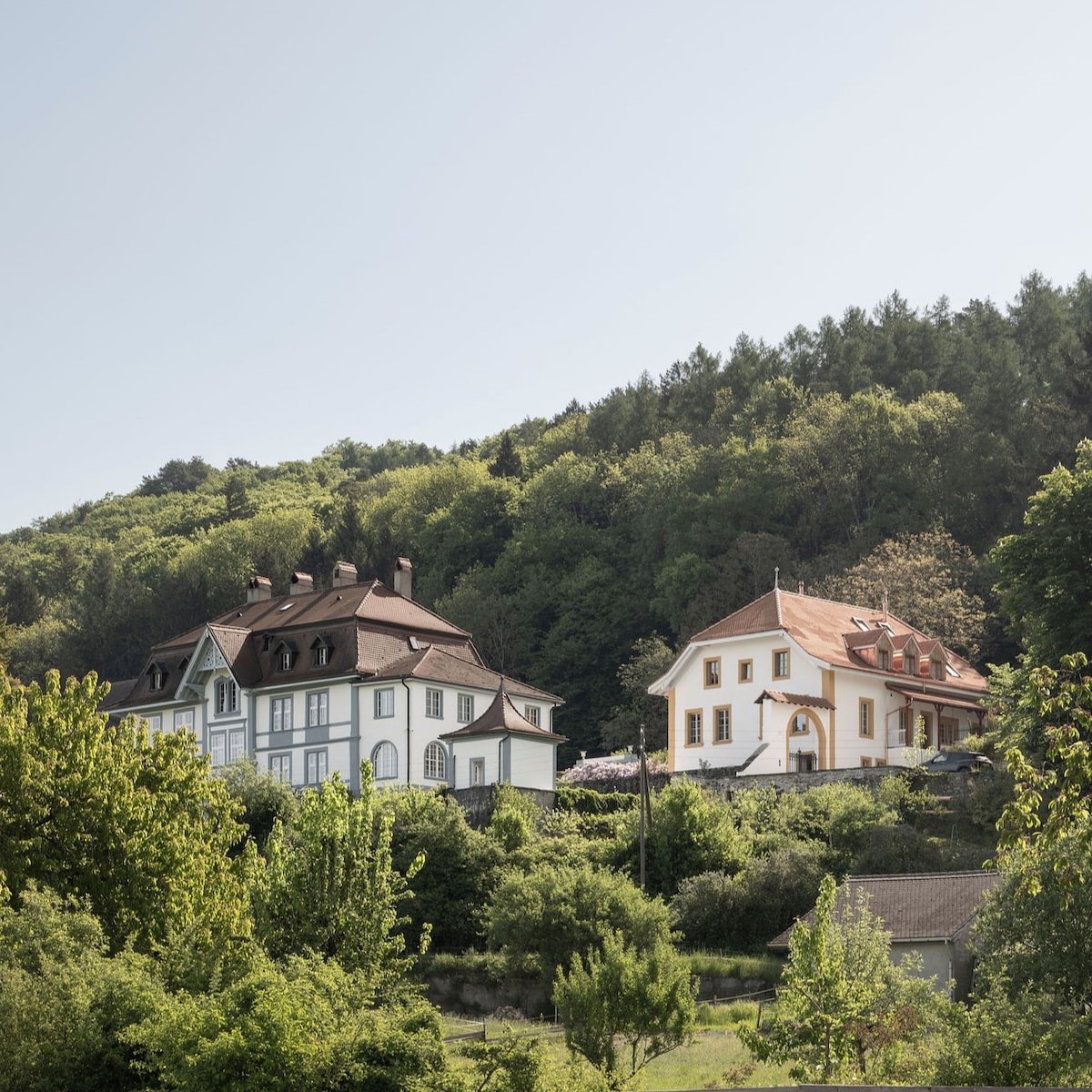 Two distinct buildings are nestled among lush greenery on a hillside. One structure features a white façade with dark trim and a sloping roof, while the other displays an orange and white exterior with multiple gabled roofs. Both offer a connection to the surrounding nature.