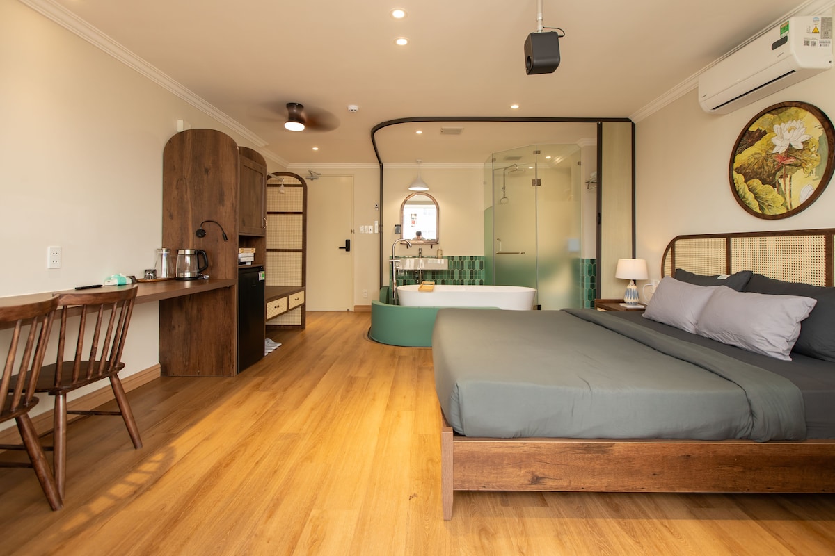 A spacious room features a large bed complemented by wood accents and warm wood flooring. A round bathtub is situated next to a glass-enclosed shower. A small dining table and chairs are arranged near the kitchen area, enhancing functionality and comfort.