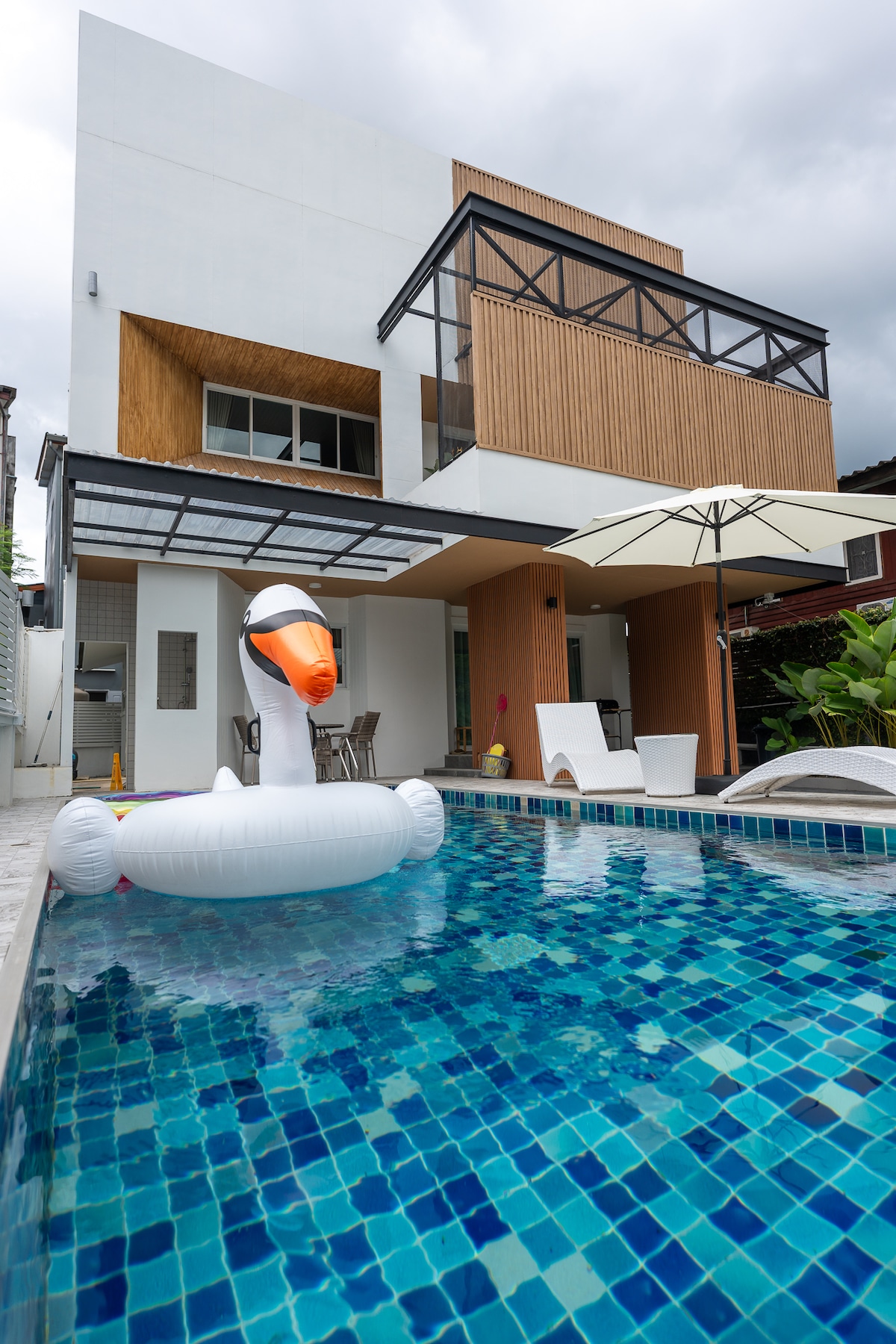 A modern villa exterior is shown, featuring a large private swimming pool with vibrant blue tiles. A white inflatable swan rests in the water alongside comfortable outdoor seating and a large umbrella for shade. The building façade is characterized by a blend of wood and sleek modern design.