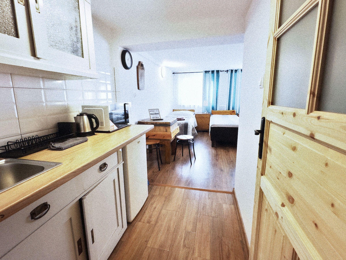 A well-lit space features a kitchenette with light wood surfaces and essential appliances. The layout includes two beds, a dining area with a wooden table, and a cozy chair. Large windows provide natural light and are dressed in soft curtains.