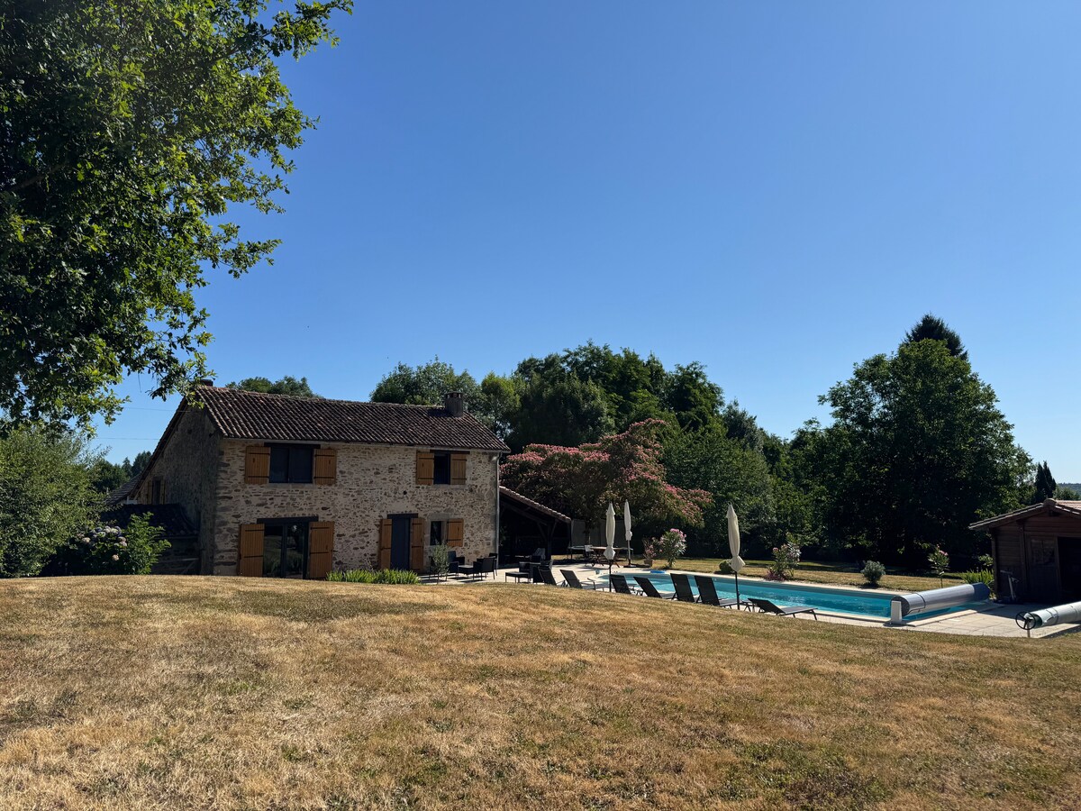 A charming stone house is framed by lush greenery, with the inviting blue of a heated swimming pool visible in the background. Sun loungers and parasols are arranged around the pool, creating a comfortable outdoor space for relaxation. The grassy area leads towards the home.