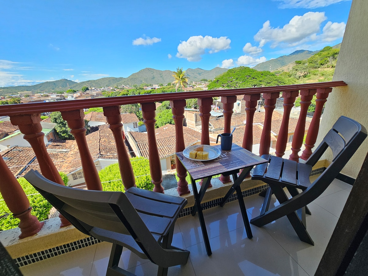 A private balcony features two wooden chairs and a small table. A refreshing drink and a plate of food are placed on the table. The serene landscape showcases green mountains and a clear blue sky, enhancing the inviting outdoor space.