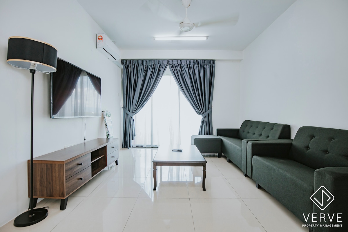 The living hall features a sleek design with a dark sofa set and a coffee table centered in the room. A smart TV is mounted on the wall, while large glass doors, covered by curtains, provide natural light and access to the outdoor area.