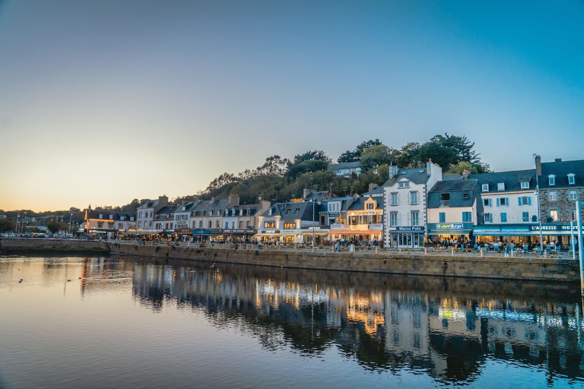 The waterfront view captures a charming row of buildings lining the harbor. As the sun sets, warm lights illuminate the façades, reflected in the calm water. Lush greenery atop the hills provides a natural backdrop, enhancing the serene atmosphere of this coastal town.