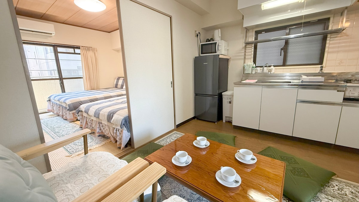 An open living space displays two double beds in the background, accented by light blue bedding. A well-equipped kitchen features modern appliances, including a refrigerator. A low wooden table is set with tea cups, situated on a soft area rug in front of a comfortable seating arrangement.