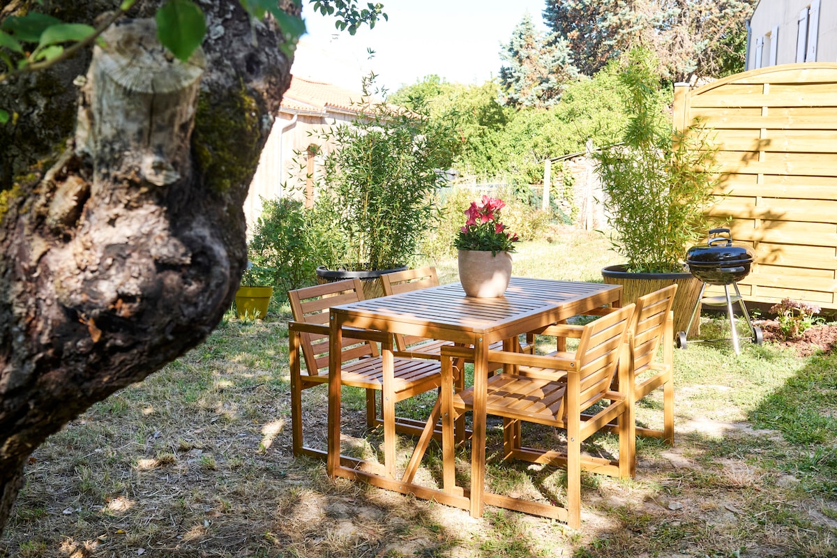 An outdoor dining area is highlighted, featuring a wooden table surrounded by six matching chairs. A planter with vibrant flowers sits at the center of the table, and a barbecue grill is positioned nearby. The green garden space is filled with natural light.