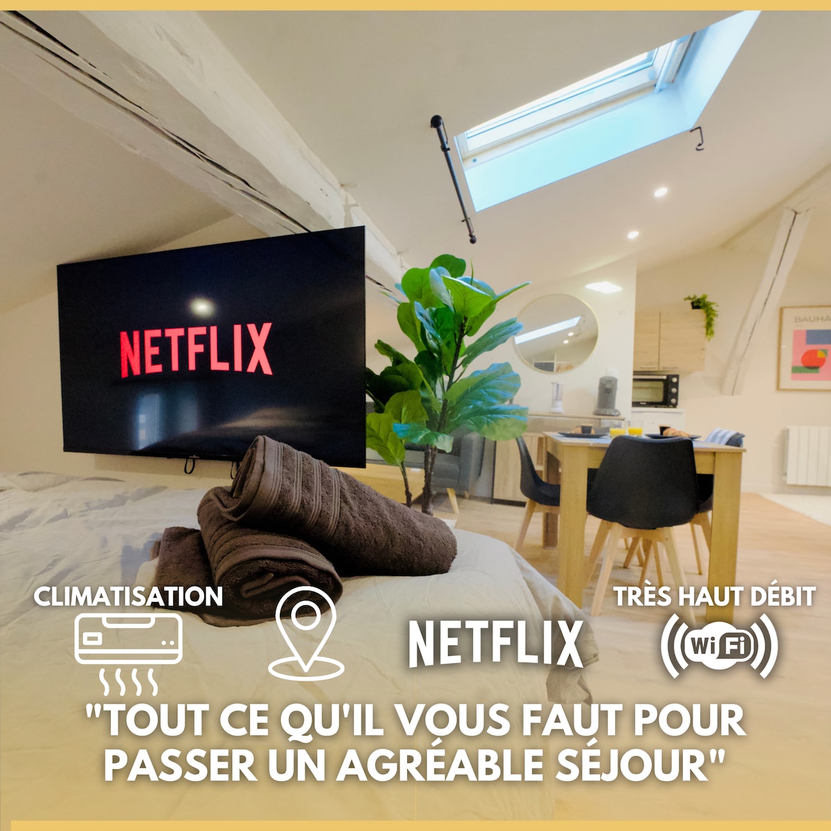The living area features a large TV displaying the Netflix logo, complemented by a cozy bed adorned with folded towels. A dining table is visible in the background, illuminated by natural light streaming through skylights. House plants add a touch of greenery to the space.