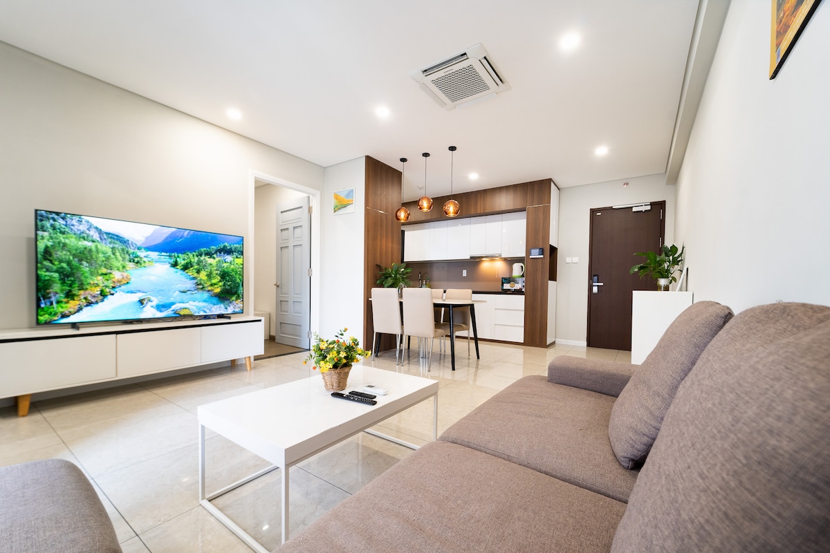 A modern living area is shown with a soft gray sofa and a white coffee table. A large smart TV is mounted on the wall, opposite a kitchen area featuring wooden cabinets and a dining space. Natural light brightens the room.