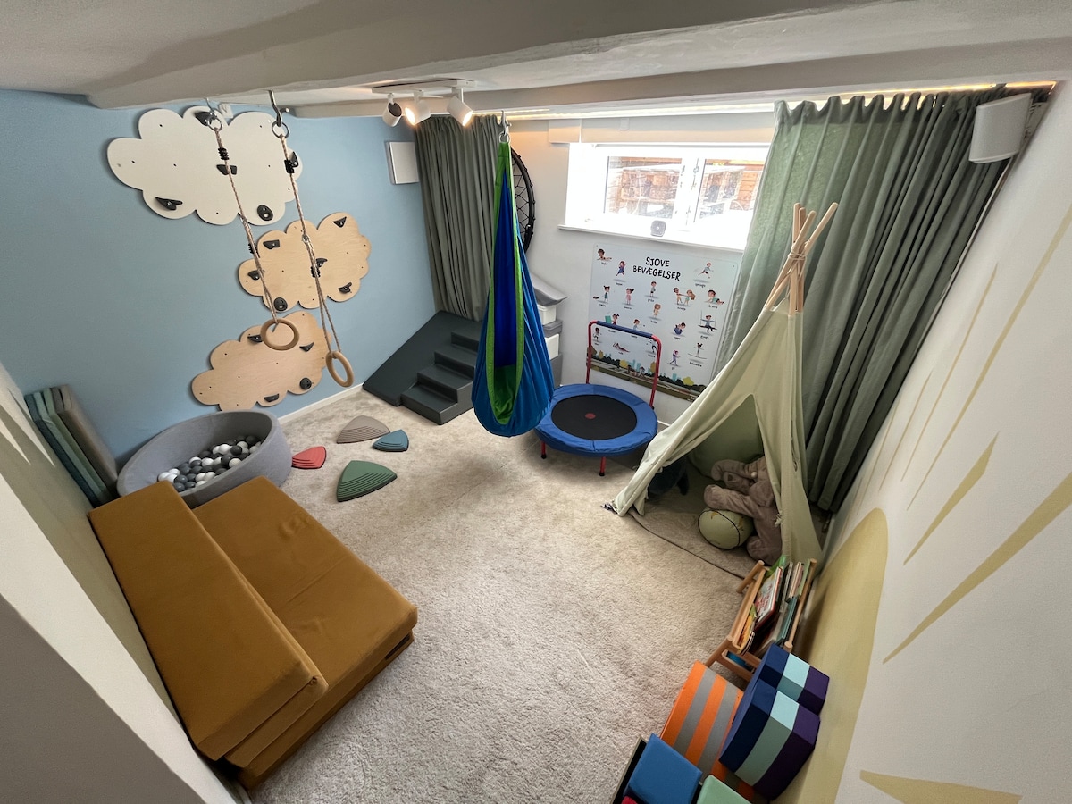 A vibrant playroom is filled with engaging features including a climbing wall, swing, and small trampoline. Soft mats are scattered on the floor, while colorful cushions provide seating. Light green curtains contribute to a cheerful atmosphere, complemented by playful wall decor and ample natural light.