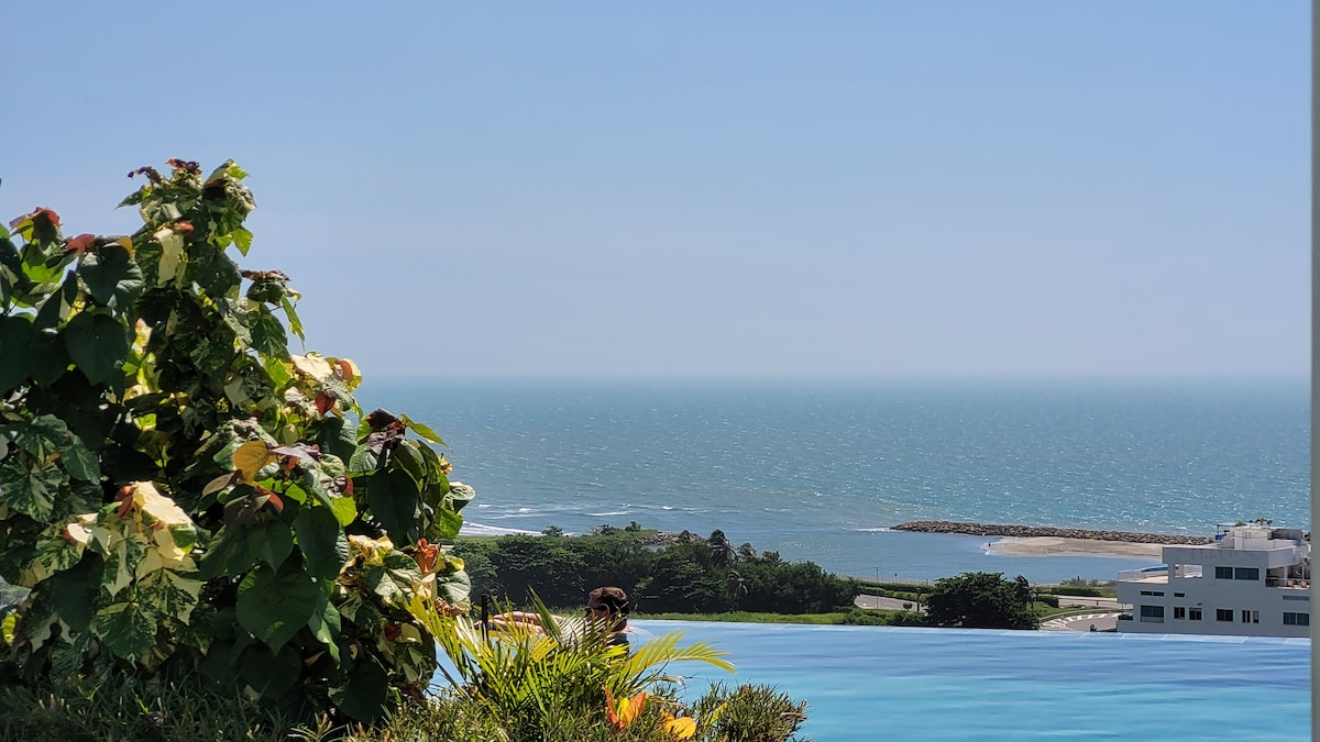A panoramic view of the ocean is displayed, with gentle waves meeting the shore. Lush green plants frame the image, contrasting with the calm blue sea and clear sky, creating a serene atmosphere that invites relaxation.