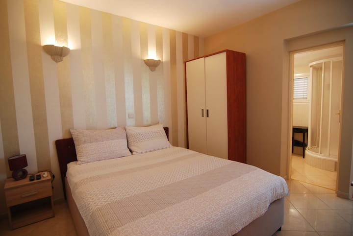 Уютна спалня с удобно двойно легло, стилно осветление, достатъчно място за съхранение и собствена баня с модерен душ. Неутралните тонове и мекото осветление създават топла, спокойна атмосфера – идеална за спокоен престой.