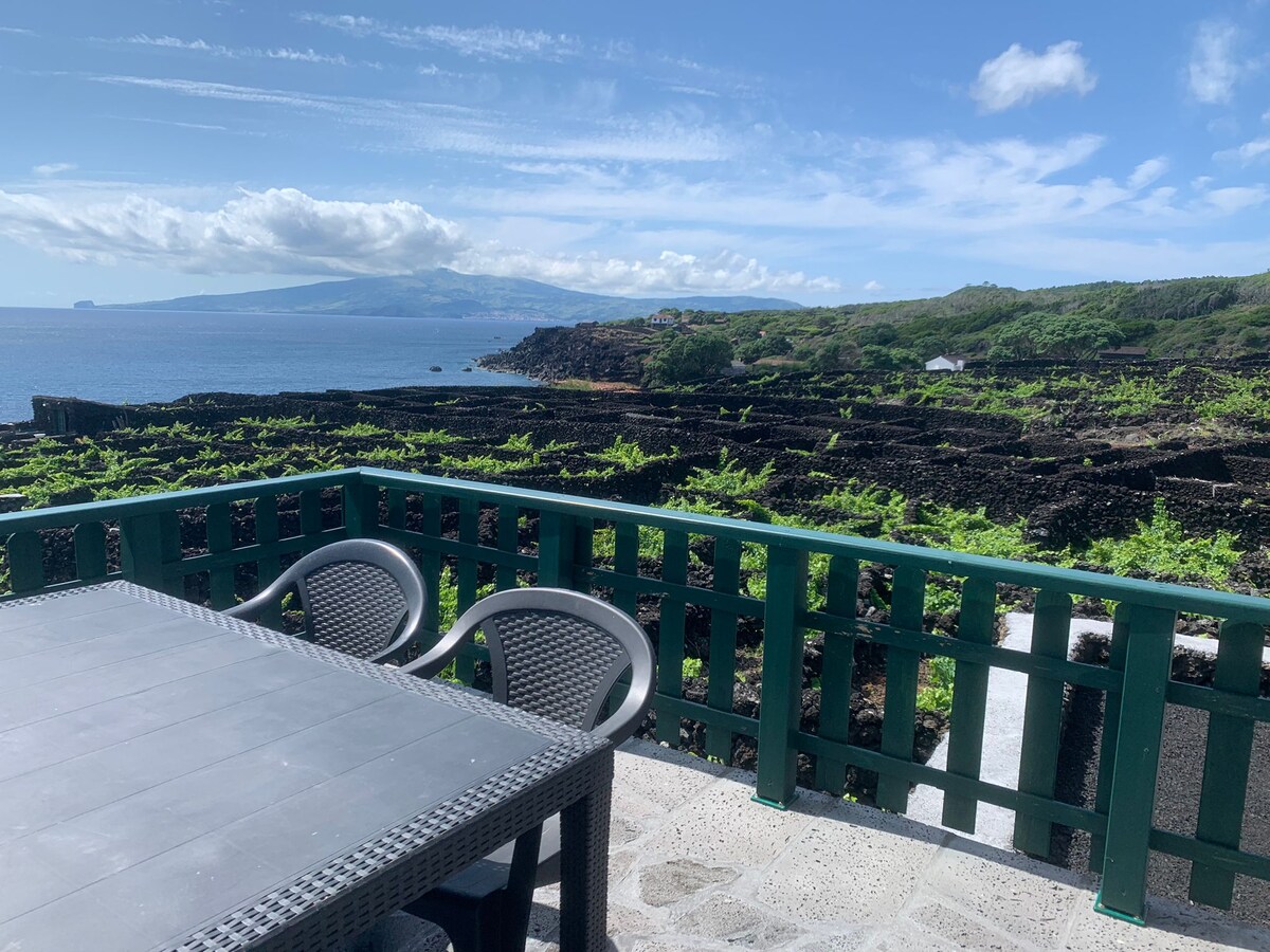 A terrace equipped with a simple dining table and four chairs offers scenic ocean views. The surrounding volcanic landscape is characterized by lush vineyards and distant mountains under a clear blue sky, creating an inviting outdoor atmosphere.
