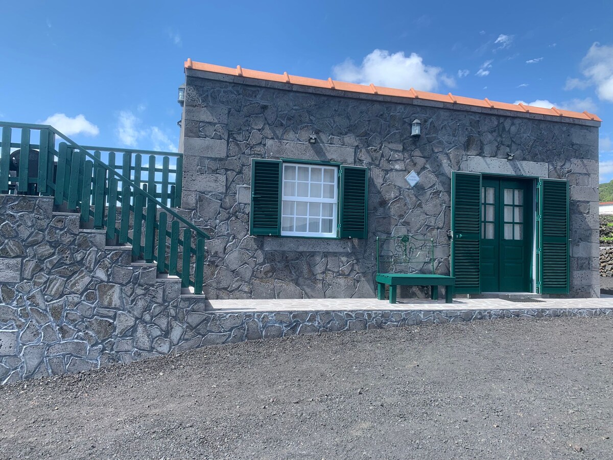 A rustic stone house features a traditional basalt facade and green wooden shutters. An inviting entrance is flanked by a charming staircase leading to a raised patio. The space is surrounded by a gravel area with coastal scenery visible in the background under a clear blue sky.