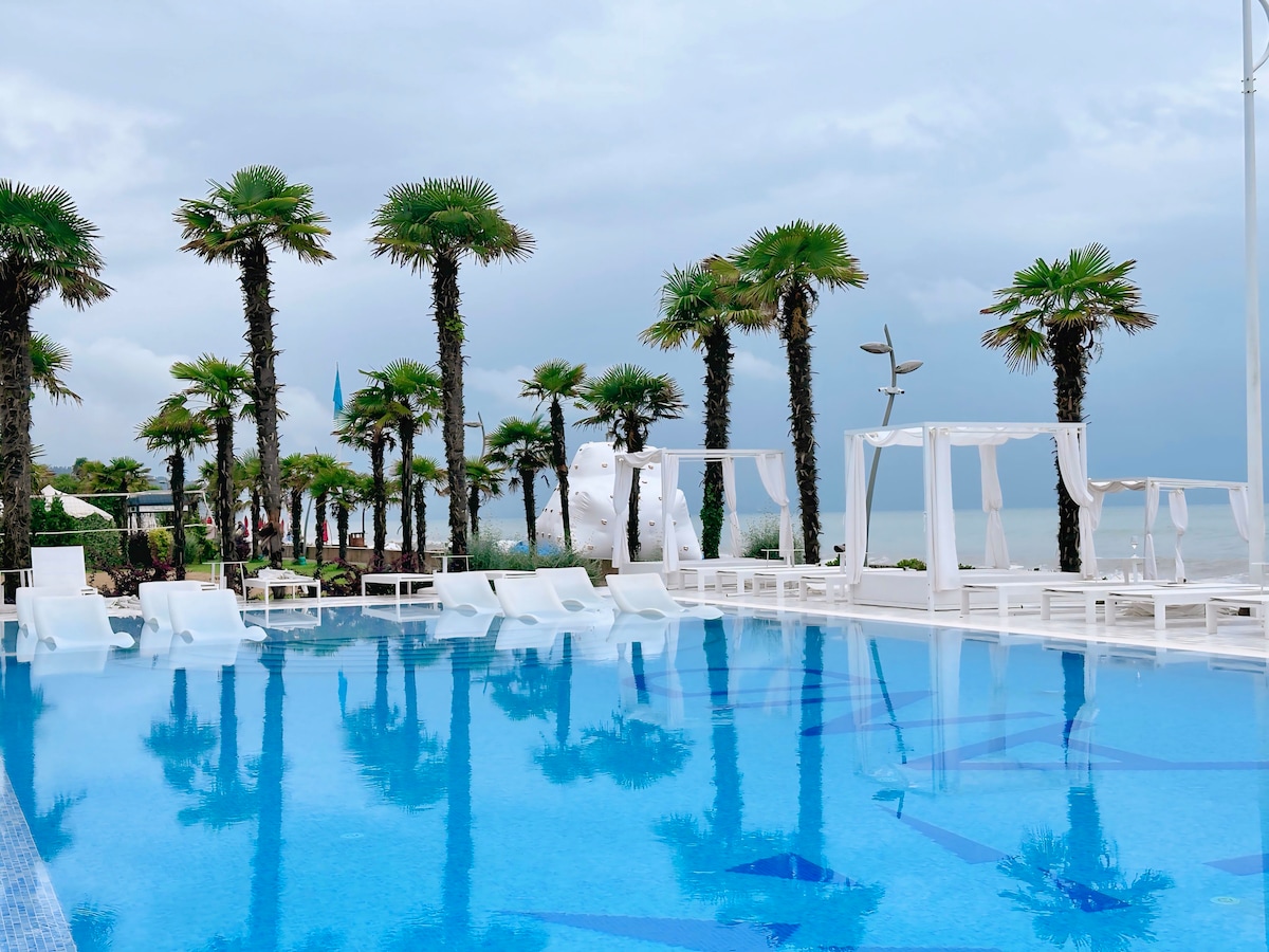 An outdoor pool area is surrounded by palm trees, featuring white lounge chairs and elegant cabanas. The pool's surface reflects the sky, with hints of turquoise and blue. Nearby, the beach is visible, contributing to a serene atmosphere.