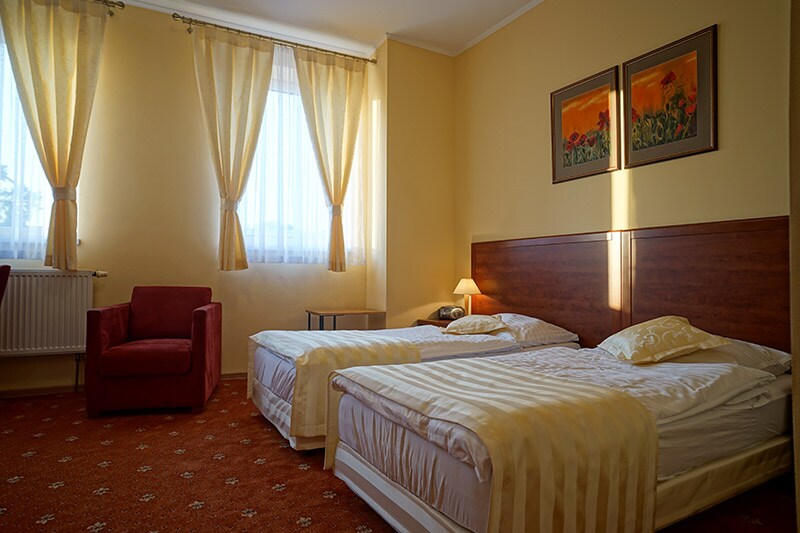 A bright and airy room features two separate beds with soft linens. Natural light is filtered through sheer curtains, illuminating the warm tones of the walls. A small table and a cozy chair are positioned near the window, complementing the space.