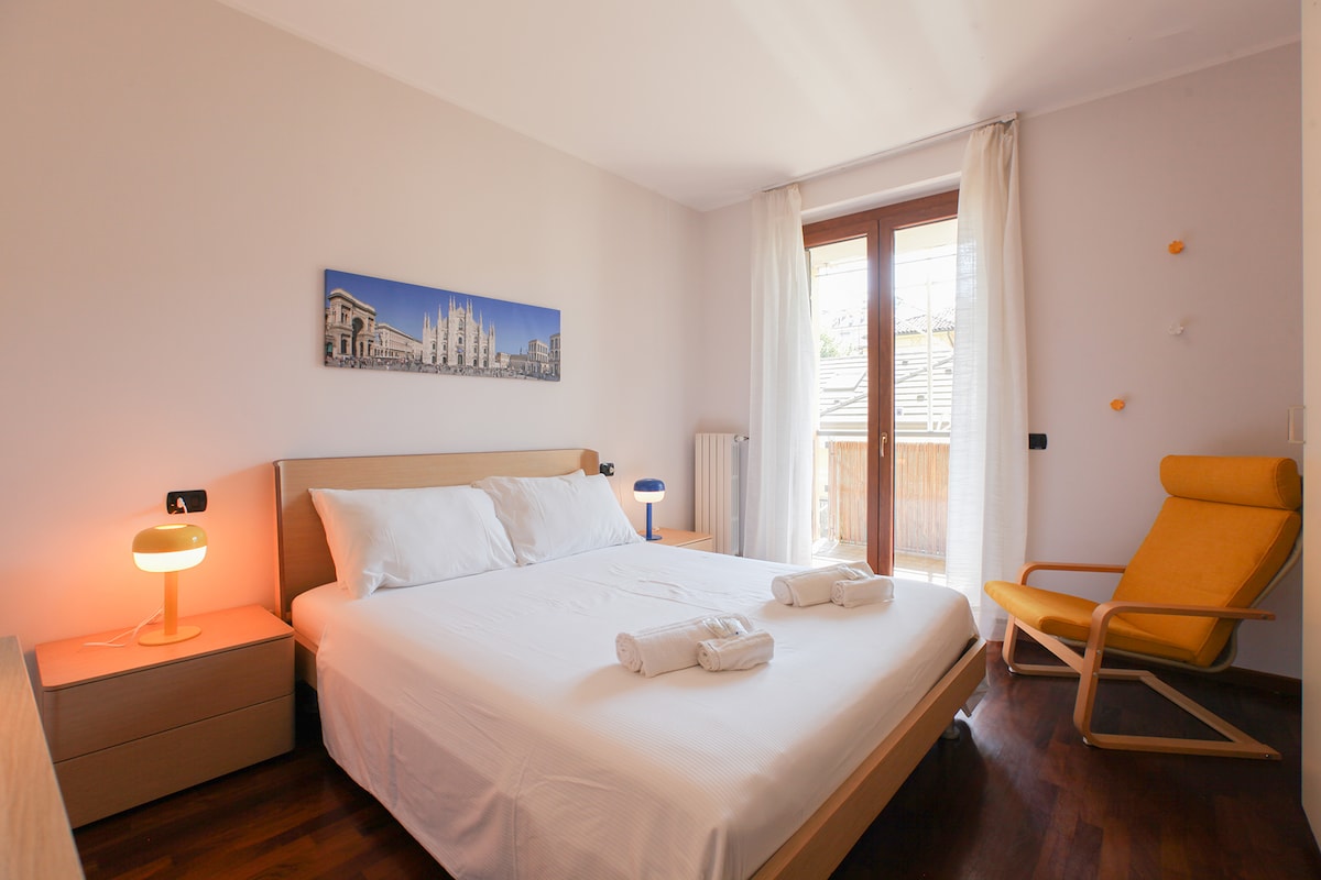 A bright bedroom features a spacious bed with neatly arranged towels, complemented by a chair in a warm tone. Natural light filters through large windows, which open to a balcony. A decorative wall piece adds a subtle artistic touch to the serene environment.