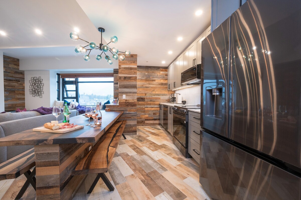 A modern kitchen and dining space is present, featuring wood-paneled walls and a sleek dining table with benches. A large refrigerator and stove are visible, alongside stylish light fixtures enhancing the ambiance. Warm natural light enters through a window, illuminating the unique flooring.