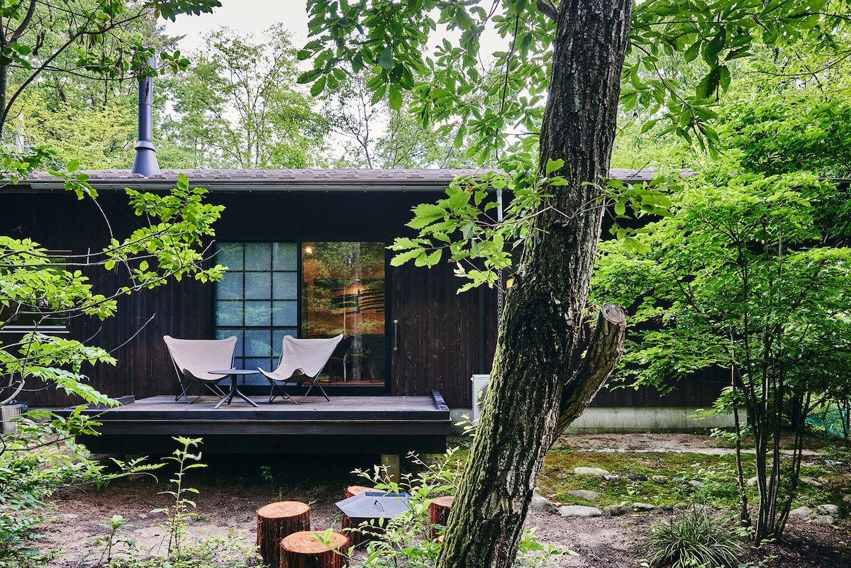 A single-story house with a dark wooden exterior is nestled among lush greenery. Large windows provide views of the surroundings. A-deck features two outdoor chairs, inviting relaxation amidst the natural setting, while wooden stumps are arranged nearby for additional seating.