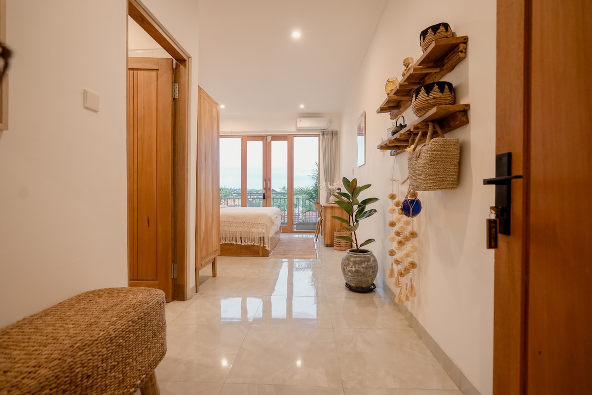 A spacious hallway is showcased with glossy tile flooring leading to the bedroom. Natural light streams through large glass doors on the right, opening to a private balcony. Decorative wall hangings and a plant add warm touches to the minimalist decor.