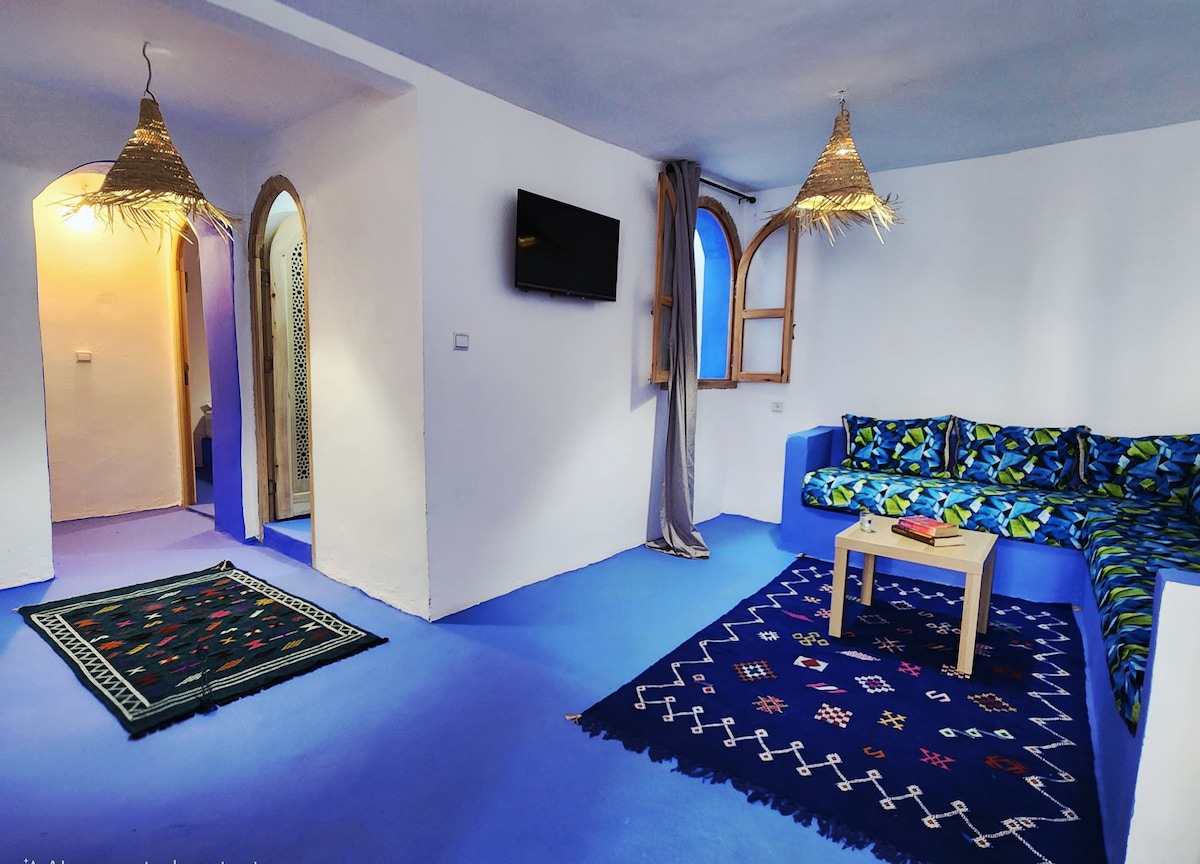 A cozy living area showcases a blue floor and white walls. A sofa adorned with vibrant floral patterns sits against the wall. Light from a nearby window enhances the space, while woven baskets hanging from the ceiling provide a decorative touch.