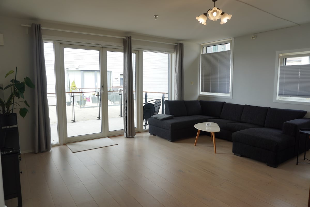 A bright living area features a large sectional sofa and a light wooden coffee table. Sliding glass doors provide access to the private balcony. Natural light fills the space through two windows adorned with light gray curtains, creating a comfortable and open environment.