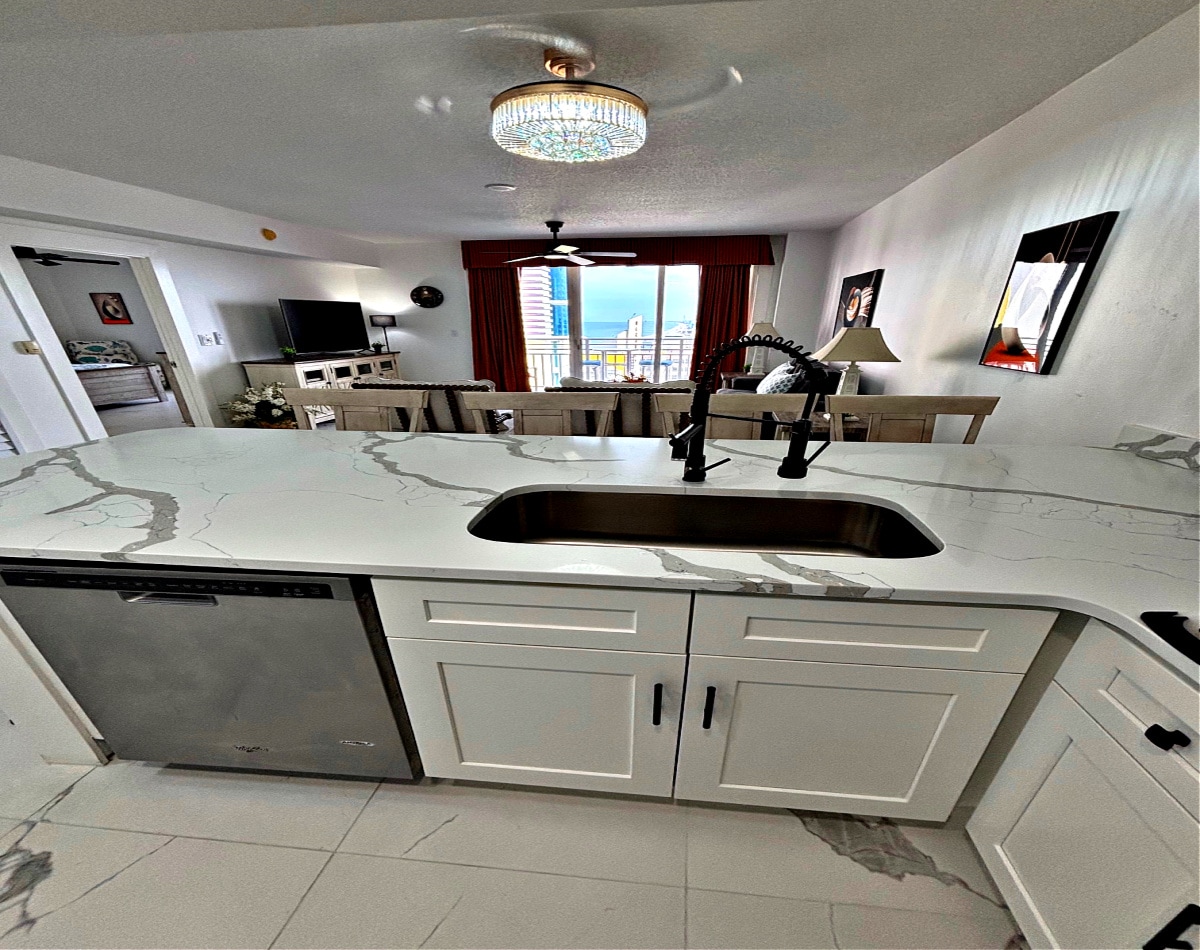 A modern kitchen area is showcased, featuring a sleek marble countertop with a double sink and dishwasher. Overhead, a chandelier adds elegant lighting. A window offers a view of the city and coastline, while a dining area is visible in the background, enhancing the open layout.